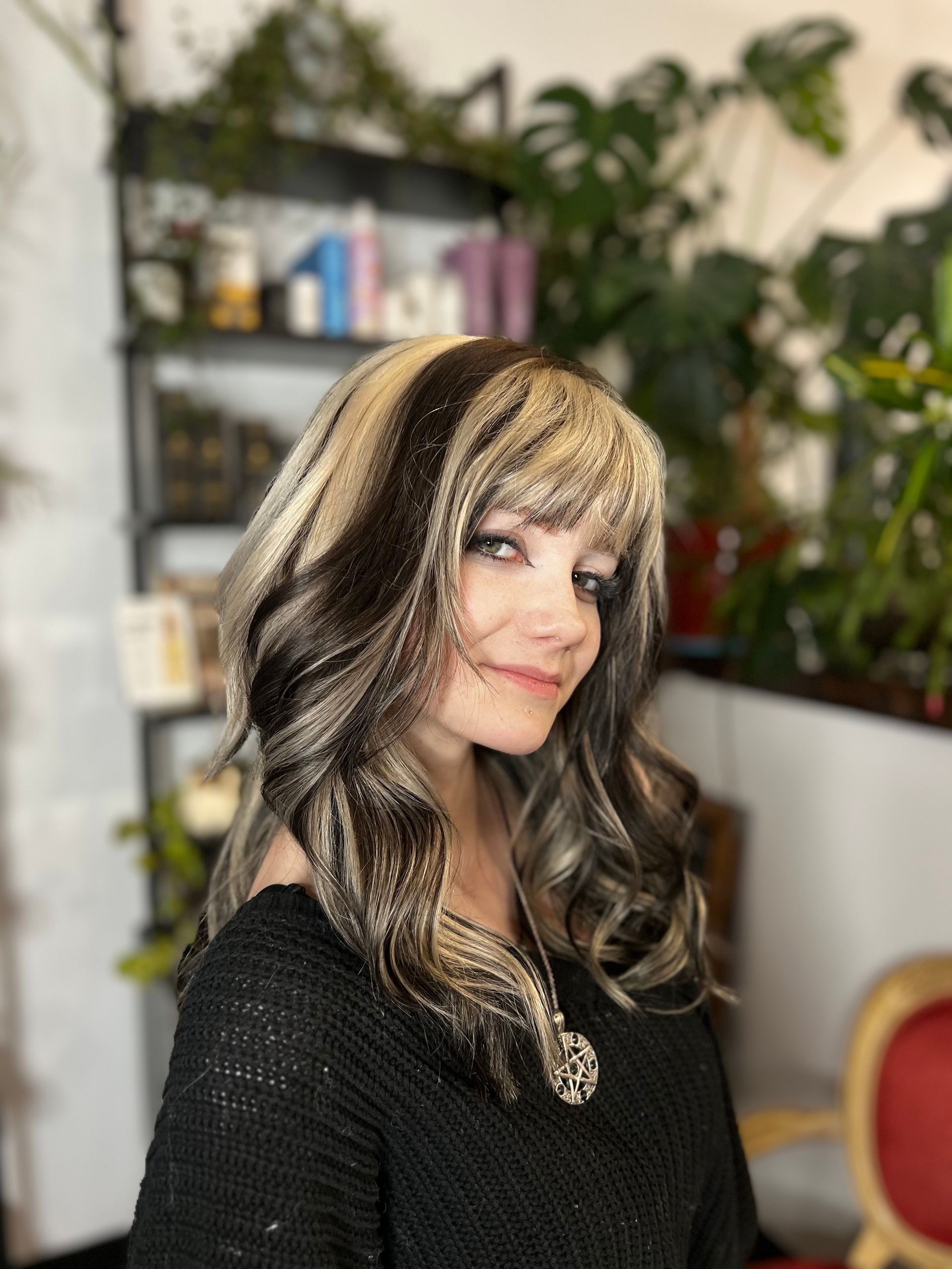 A woman wearing a black sweater and a wig is smiling in a room.