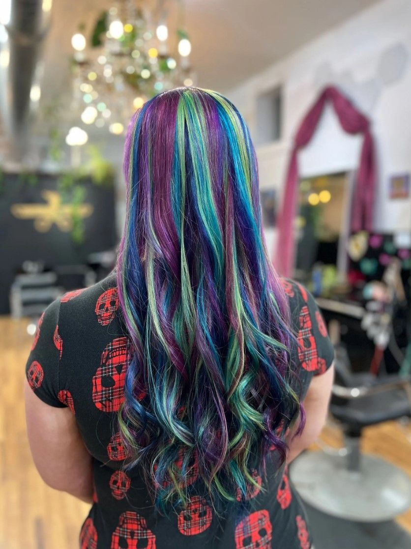 Person with long hair dyed in vibrant streaks of purple, teal, and green, in a salon setting.