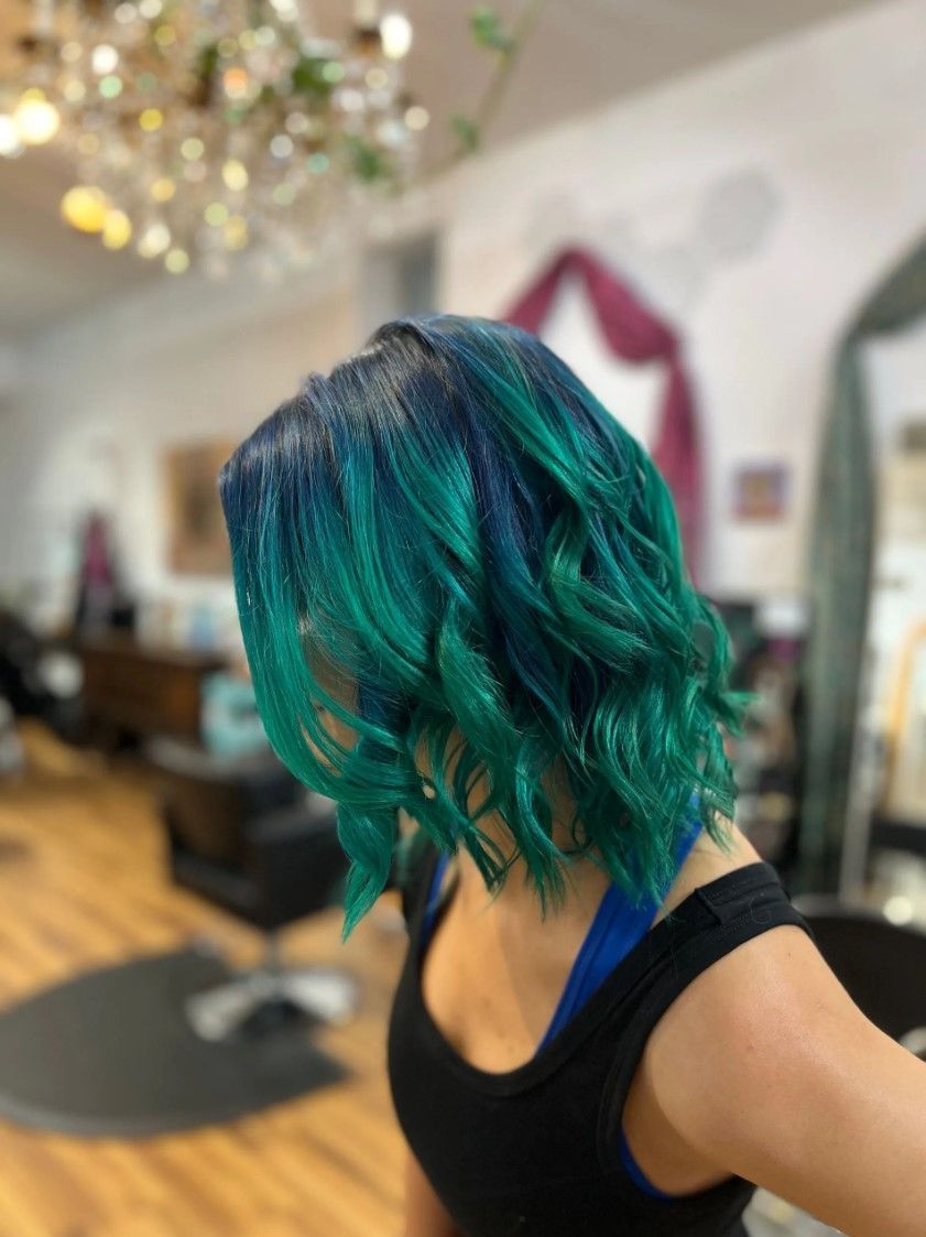 Woman with blue and teal hair in a salon, wearing a black top.