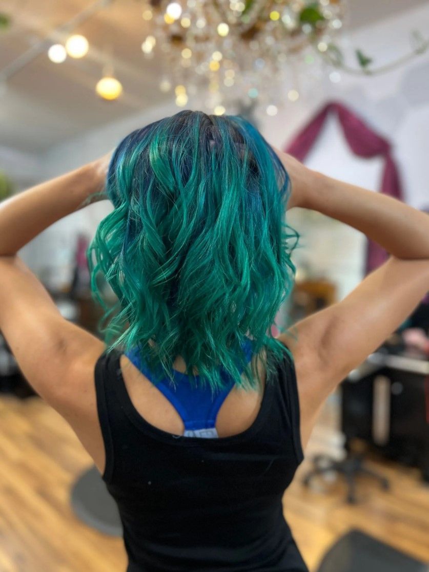 Woman with teal and green wavy hair, wearing a black tank top, in a salon.