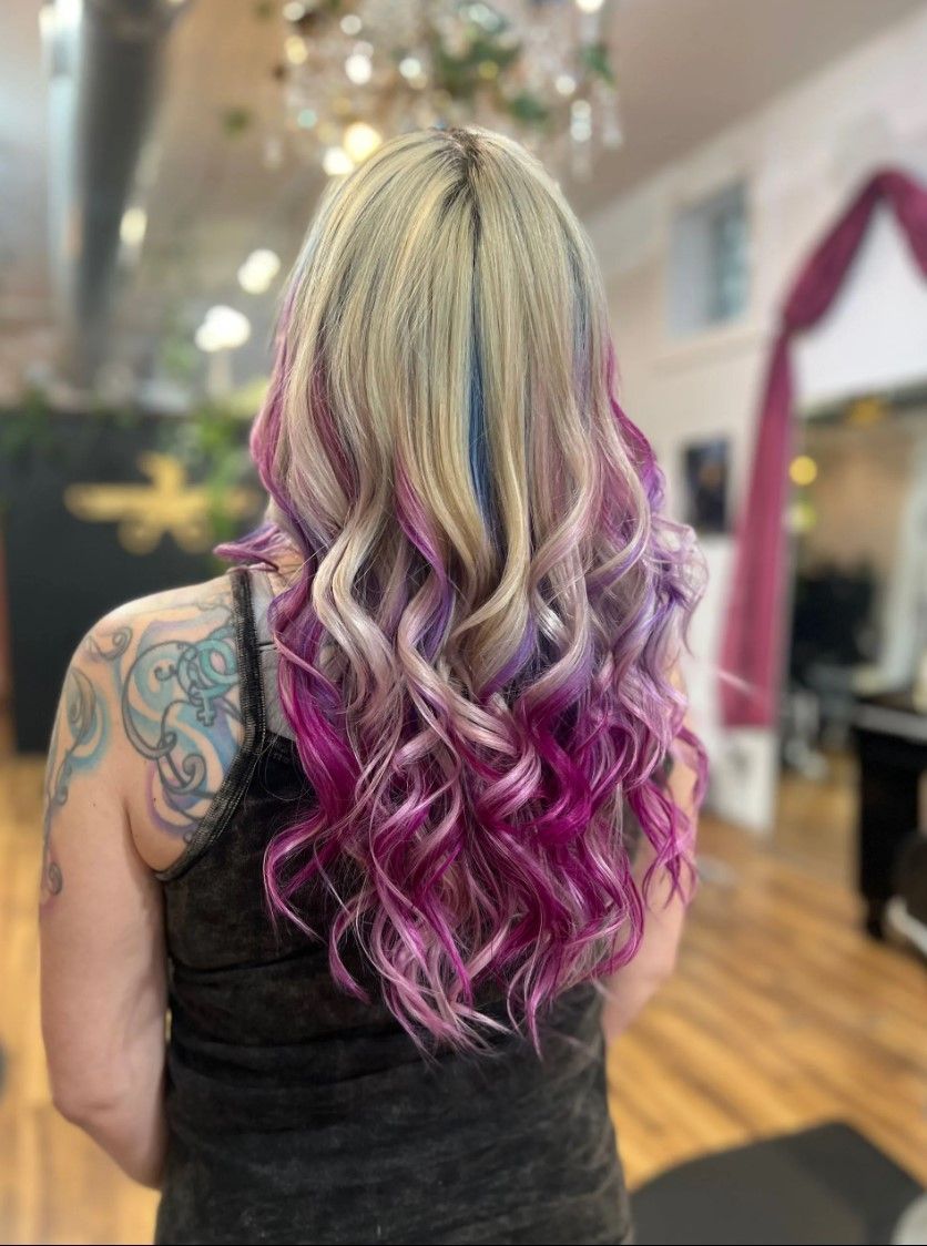 Woman with long, wavy blonde hair with pink, purple, and blue highlights, standing indoors.