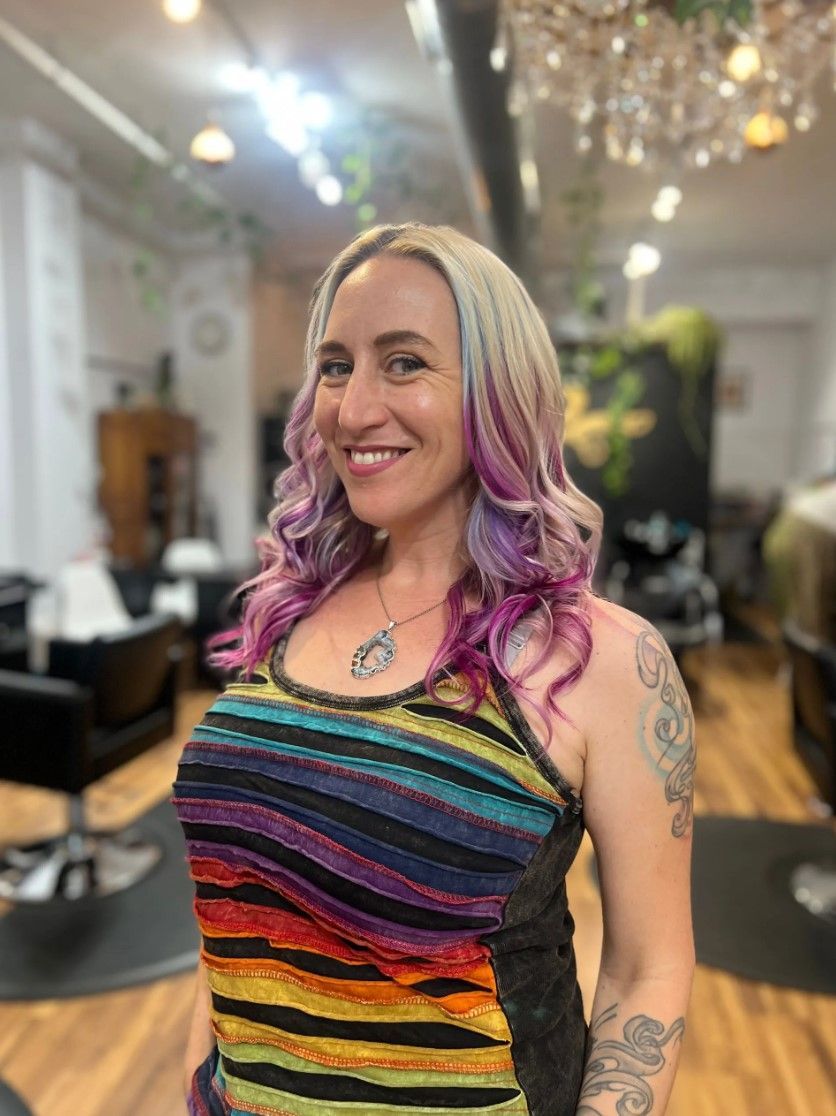 Woman with purple-streaked hair smiles, wearing a rainbow top in a salon.