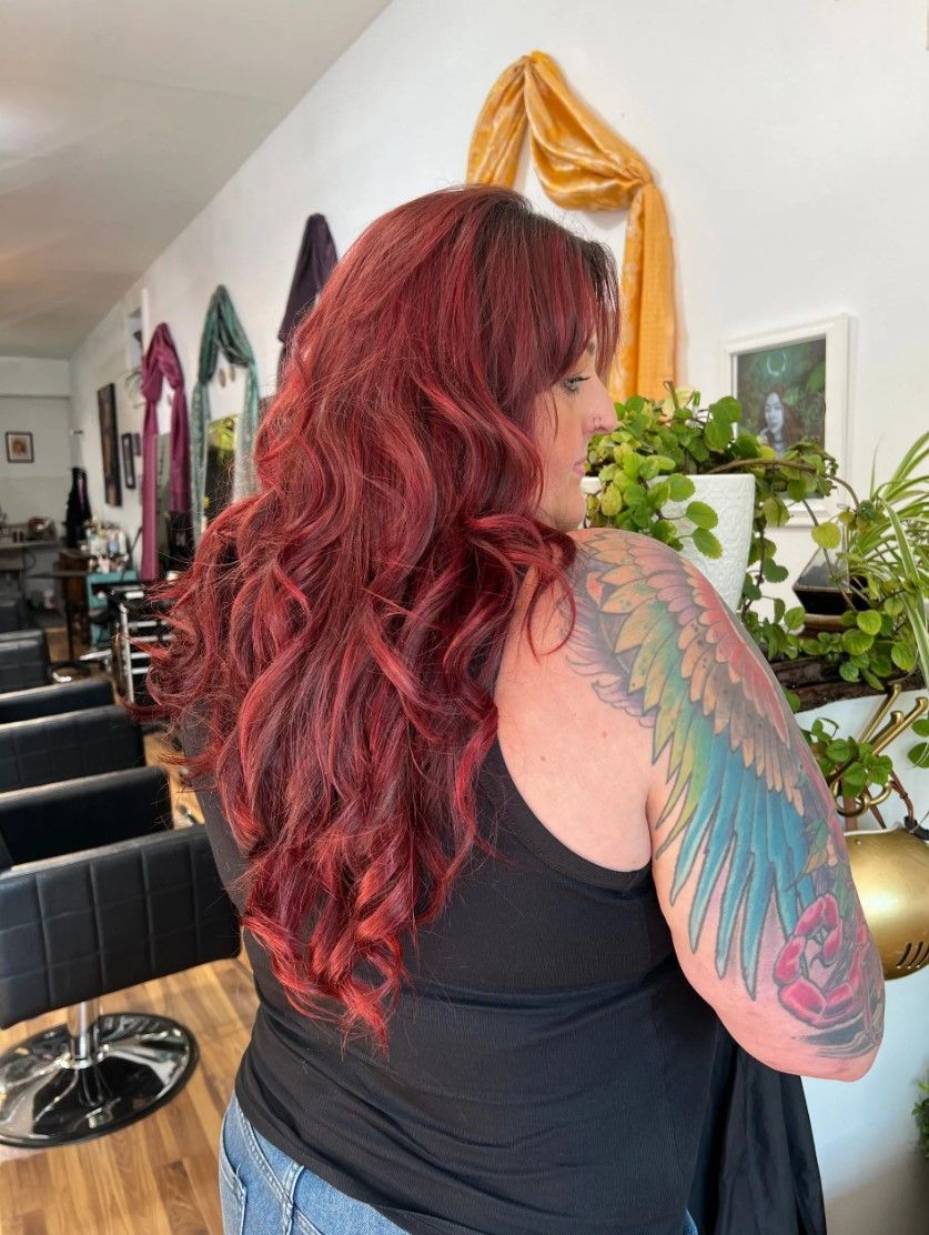 Woman with long, wavy red hair and a colorful arm tattoo; standing inside a salon.