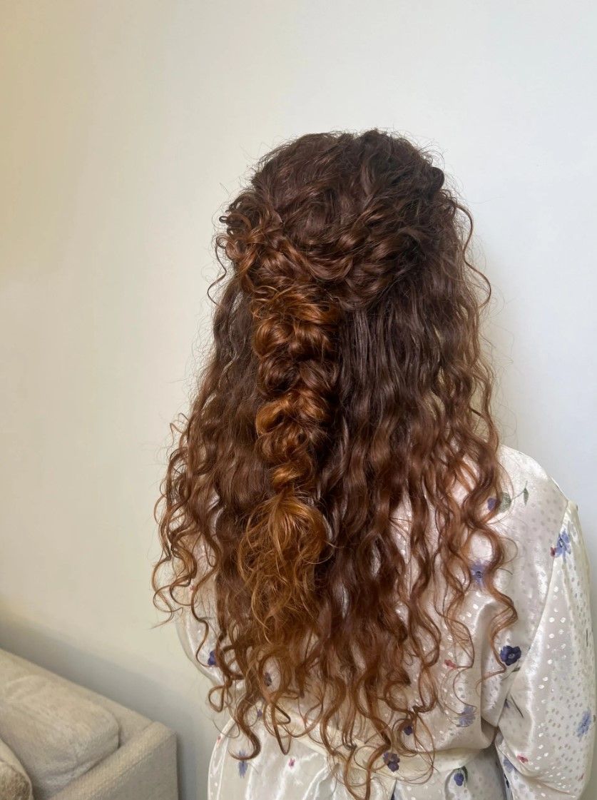 Person with long, curly, reddish-brown hair in a partial braid, standing in front of a white wall.