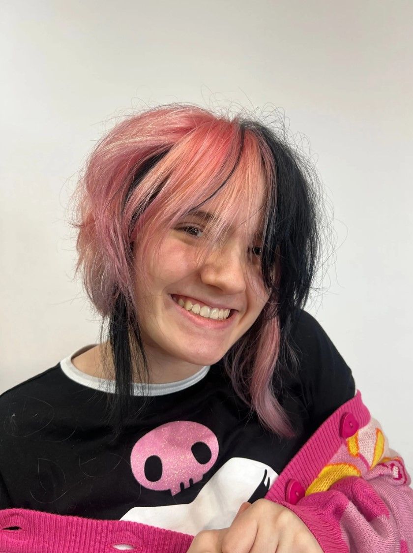 Person with pink and black hair smiles, wearing a black shirt and pink cardigan.