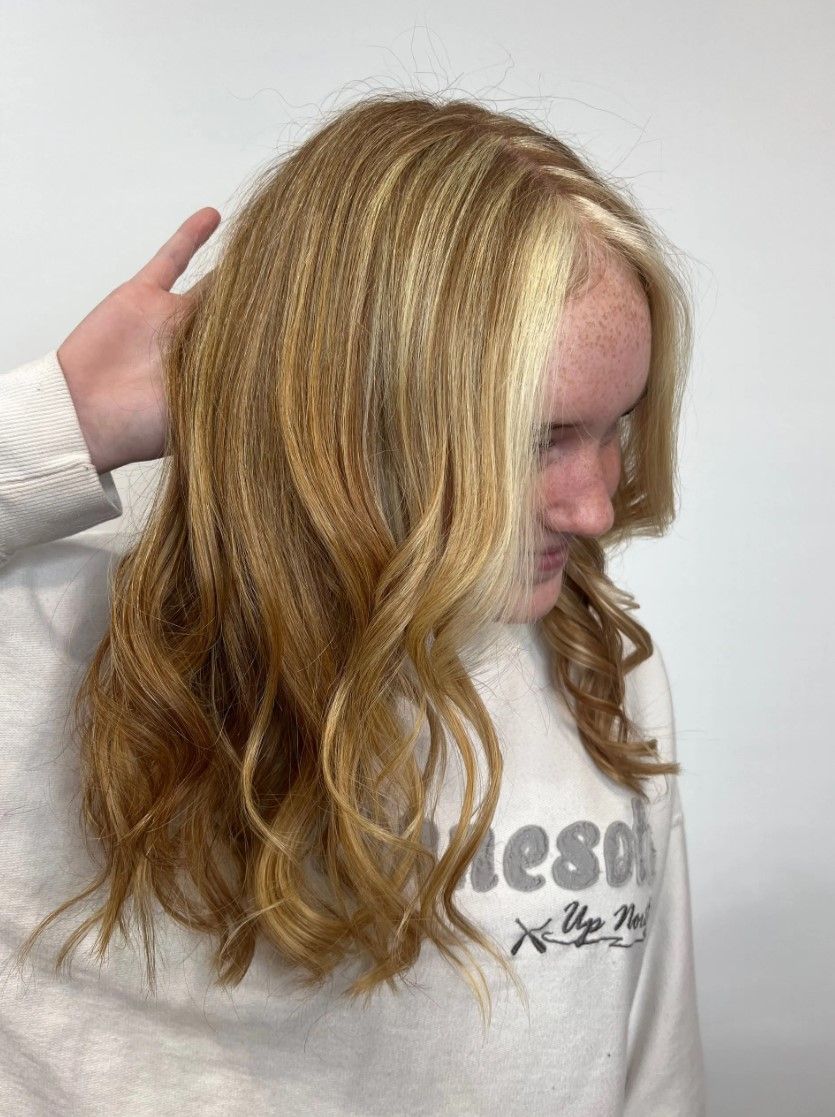 Person with blonde and brown wavy hair, wearing a white sweatshirt, hand in hair, leaning toward the right.