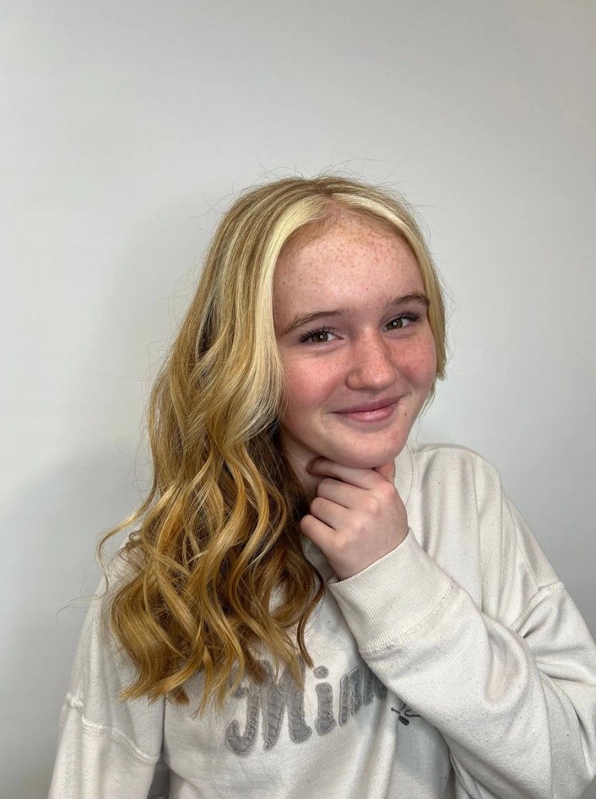 Blond-haired person with wavy hair, freckles, smiling and touching their chin, against a white background.
