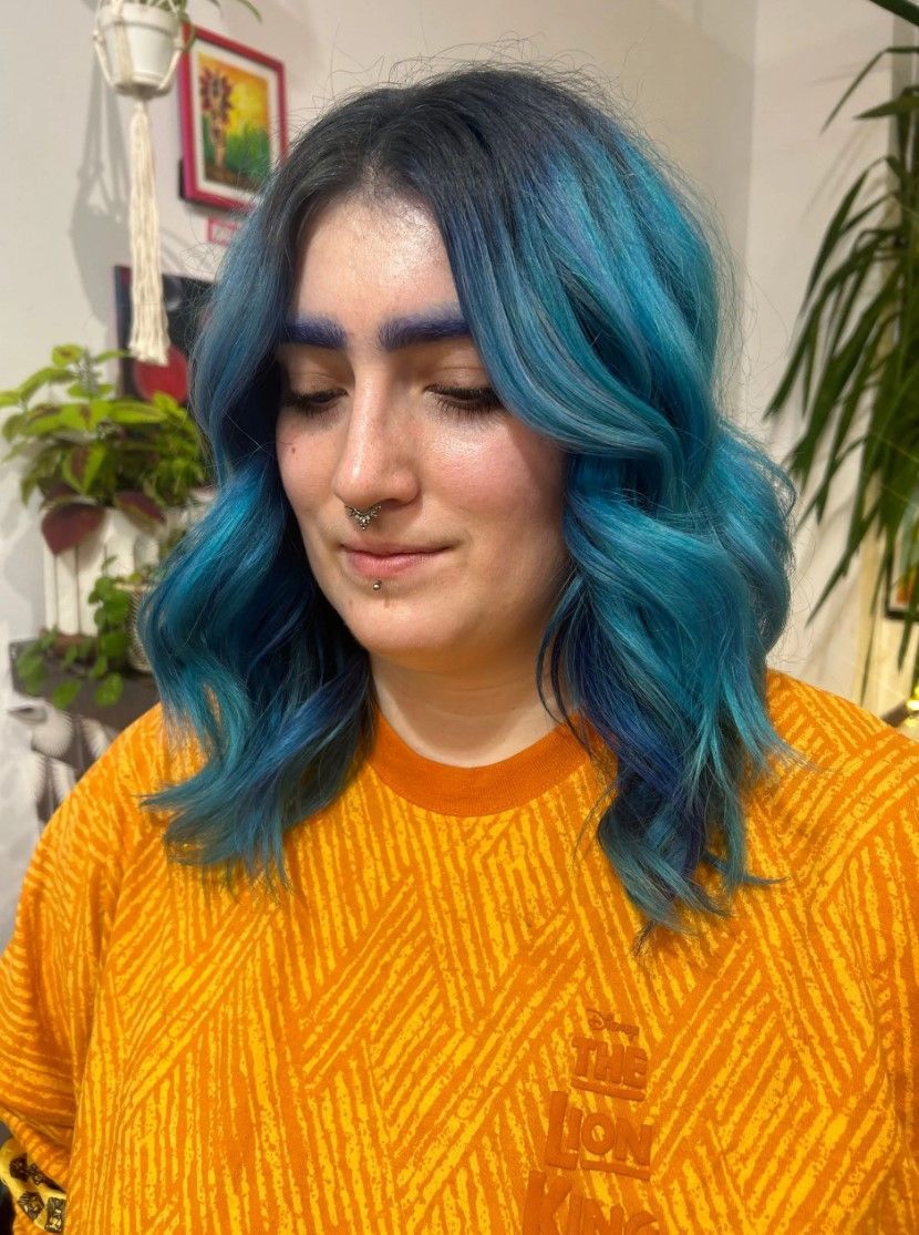 Woman with blue hair and orange shirt, looking down, in a room with plants.