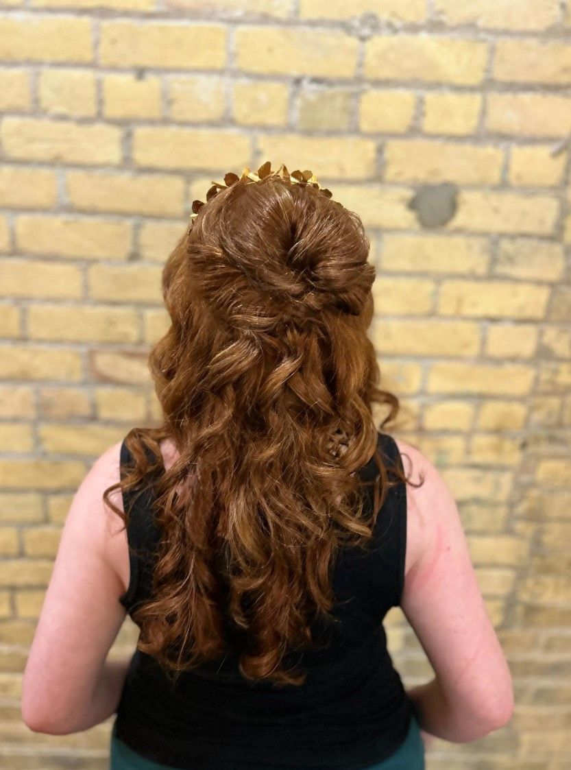 Woman with auburn hair in a half-up, half-down style, wearing a gold hairpiece, against a yellow brick wall.