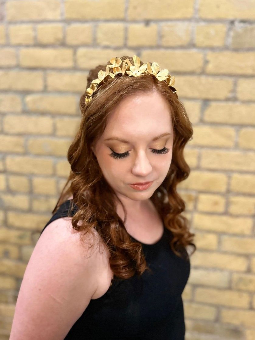 Woman with red hair wearing a gold leaf crown, looking down. Brick wall background.