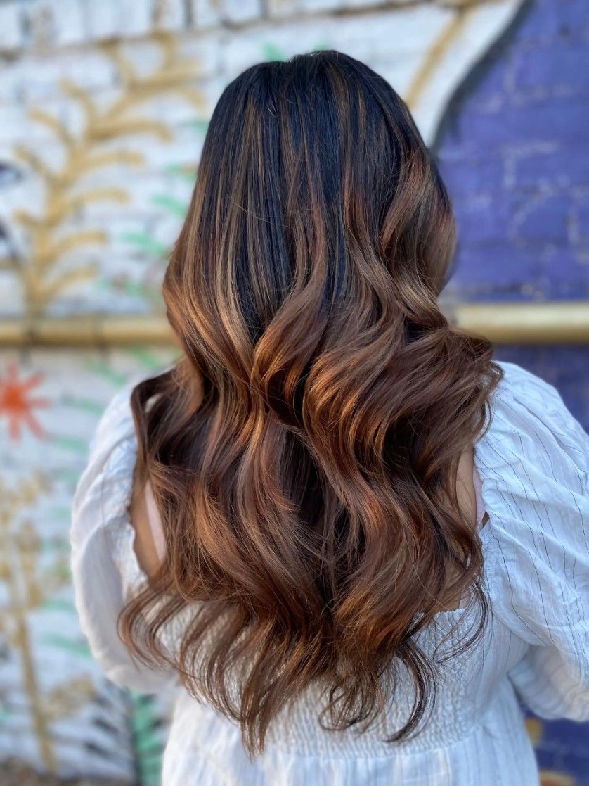 Woman with long, wavy brown hair with highlights, standing outdoors in front of a colorful wall.
