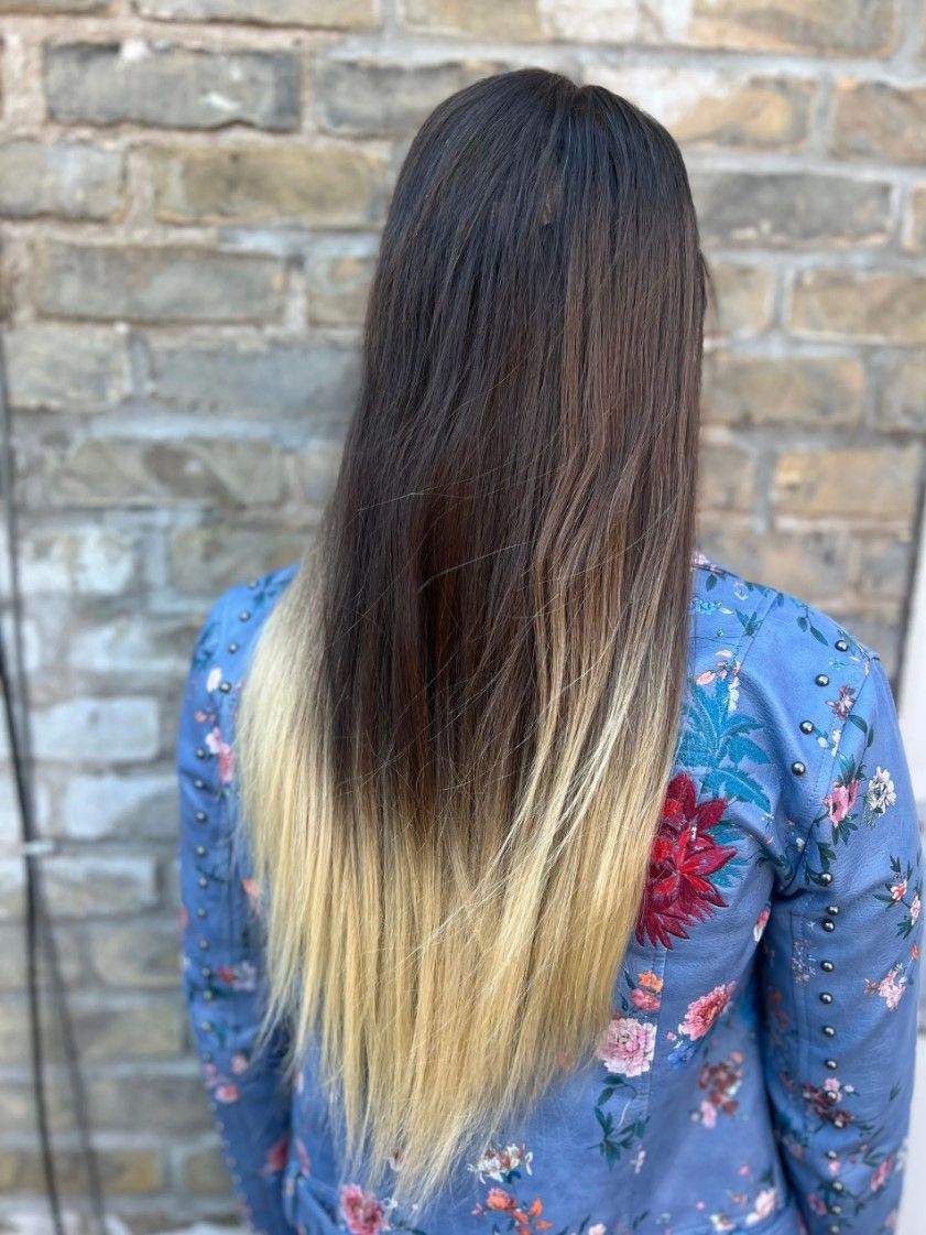 Woman with long hair featuring a dark to blonde ombre effect, wearing a blue floral shirt, standing against a brick wall.