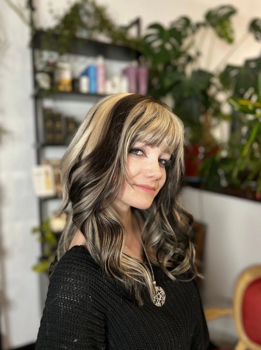Woman with dark and blonde highlighted hair smiling; indoor setting with plants and shelves.