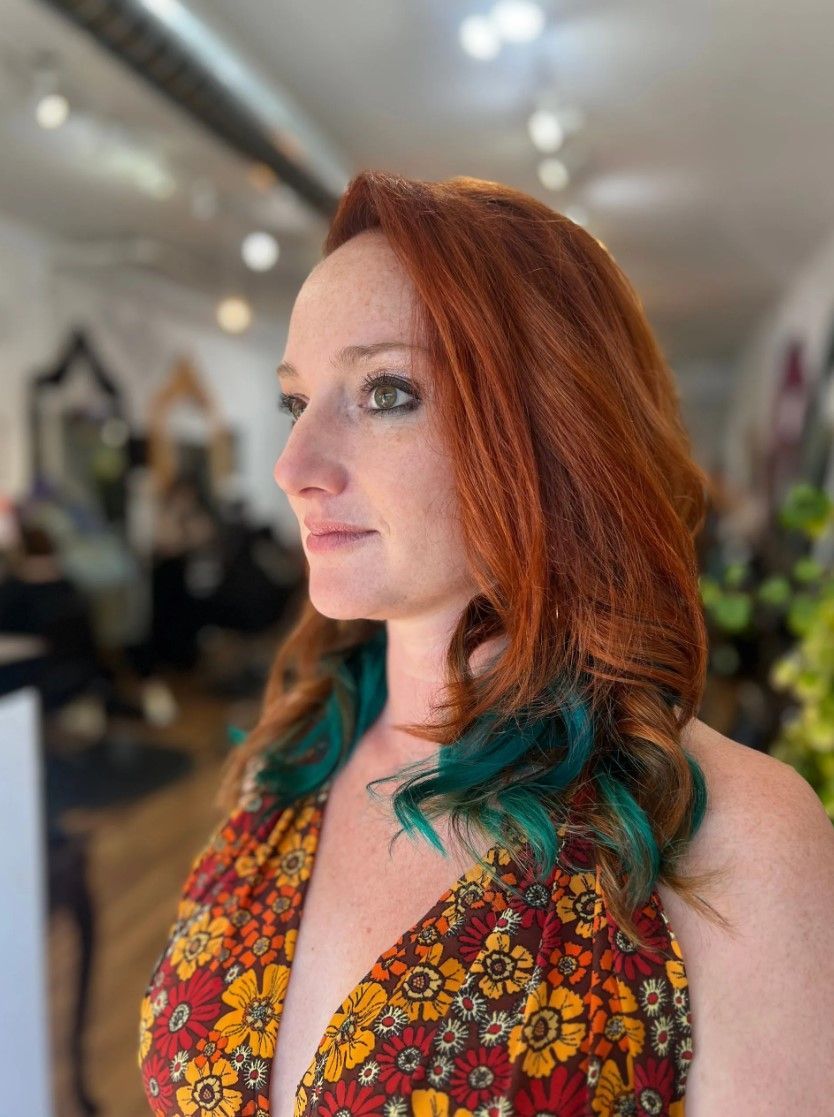 Woman with red hair and teal tips, in a salon, wearing a floral dress, looking to the side.