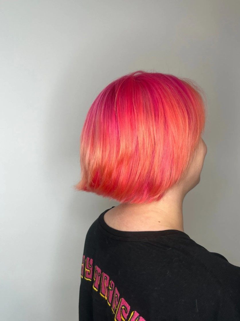 Person with short pink and orange hair, wearing a black shirt, posing against a white wall.