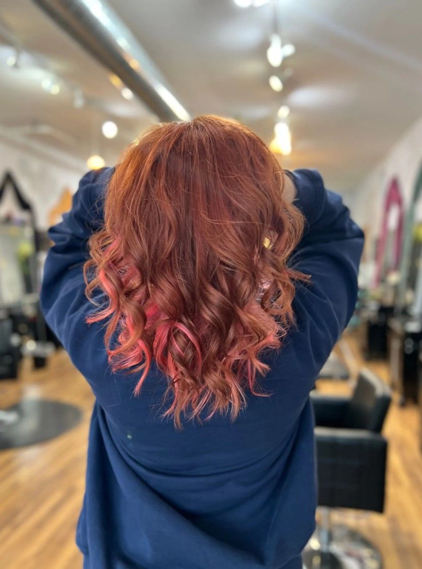 Person with curly red hair, some pink highlights, wearing a blue sweater, in a salon.