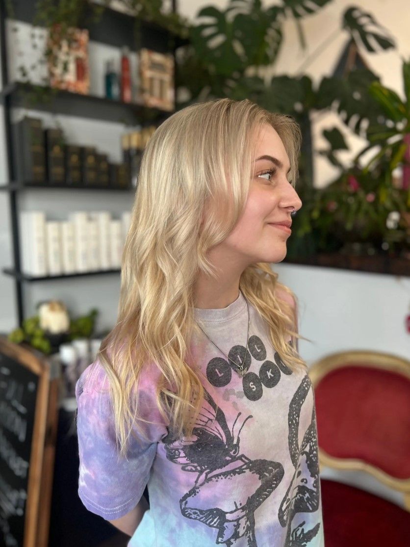 Blonde-haired person smiles, posing at a salon. They wear a tie-dye shirt with a snake design. Red chair in background.