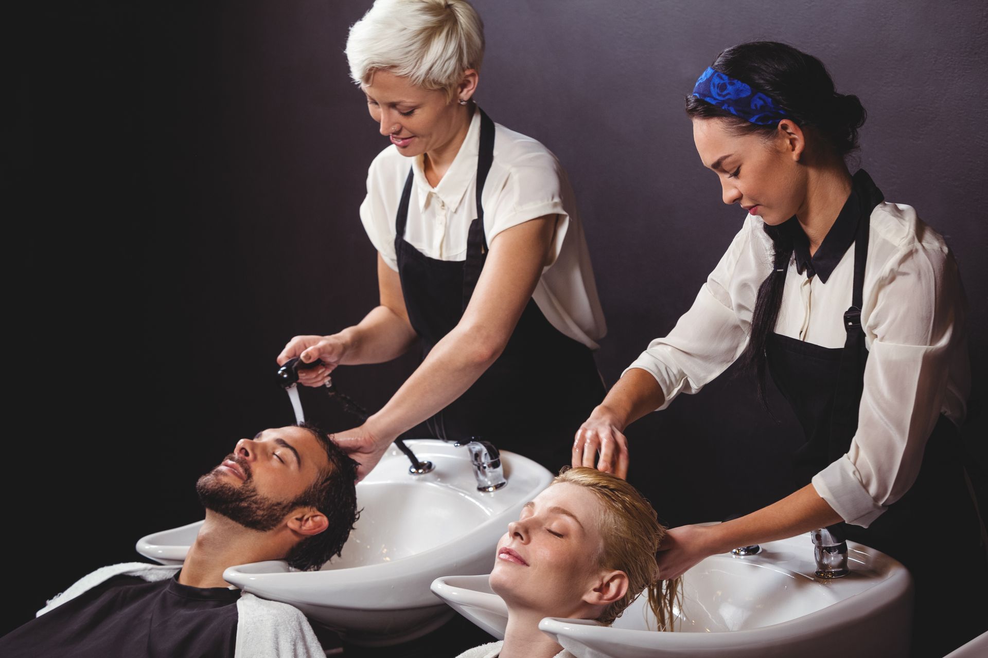 Two hairdressers washing clients' hair in a salon. Black aprons, white sinks, dark background.