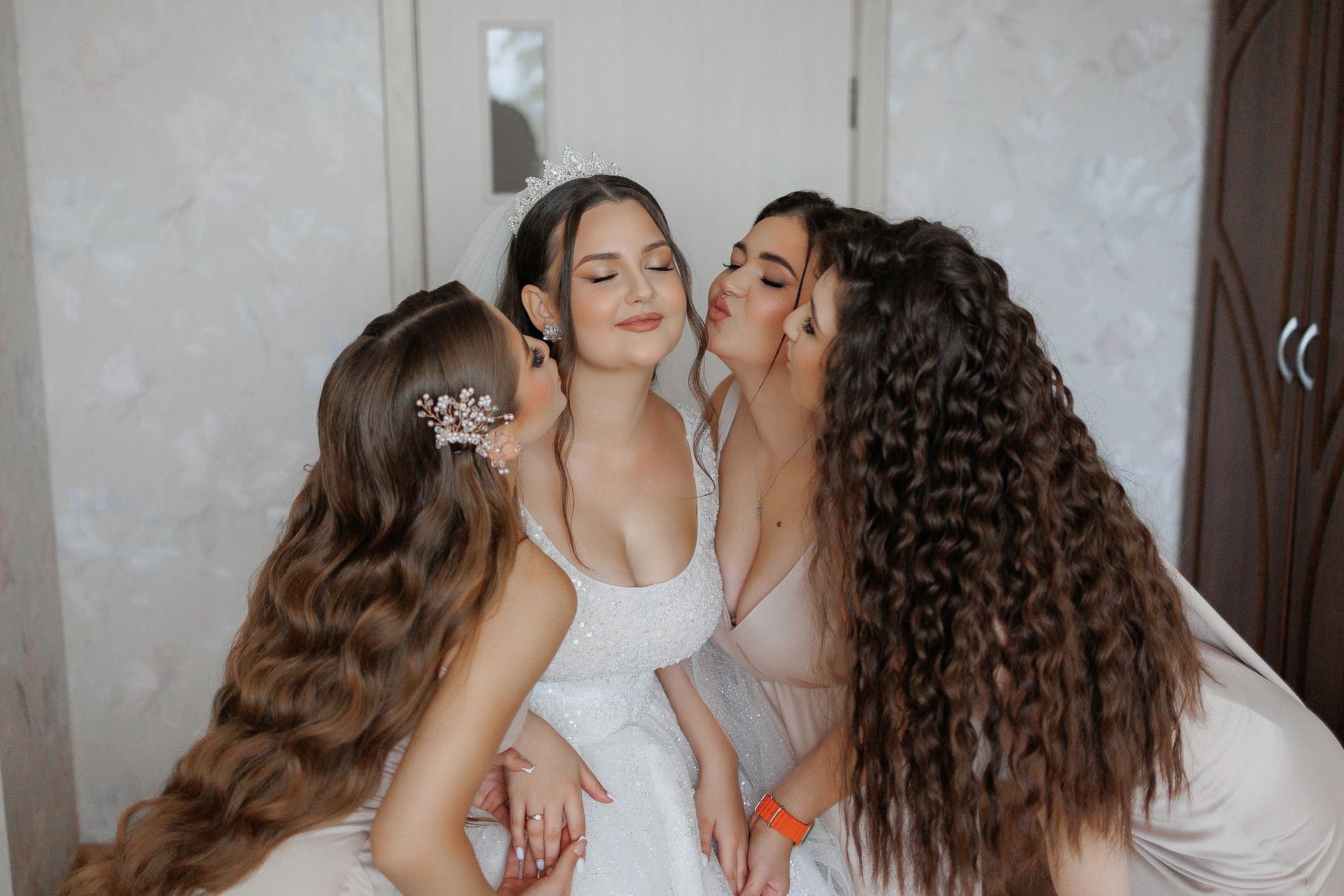 Bride surrounded by bridesmaids, getting kissed on the cheeks. Bride wears white gown, bridesmaids wear pink. Interior shot.
