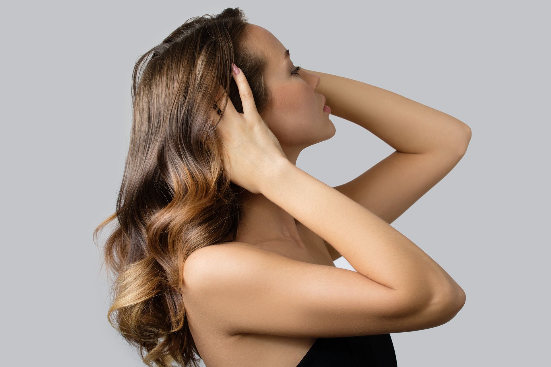 Woman with wavy brown hair, hands in hair, head turned to the side, against a gray background.