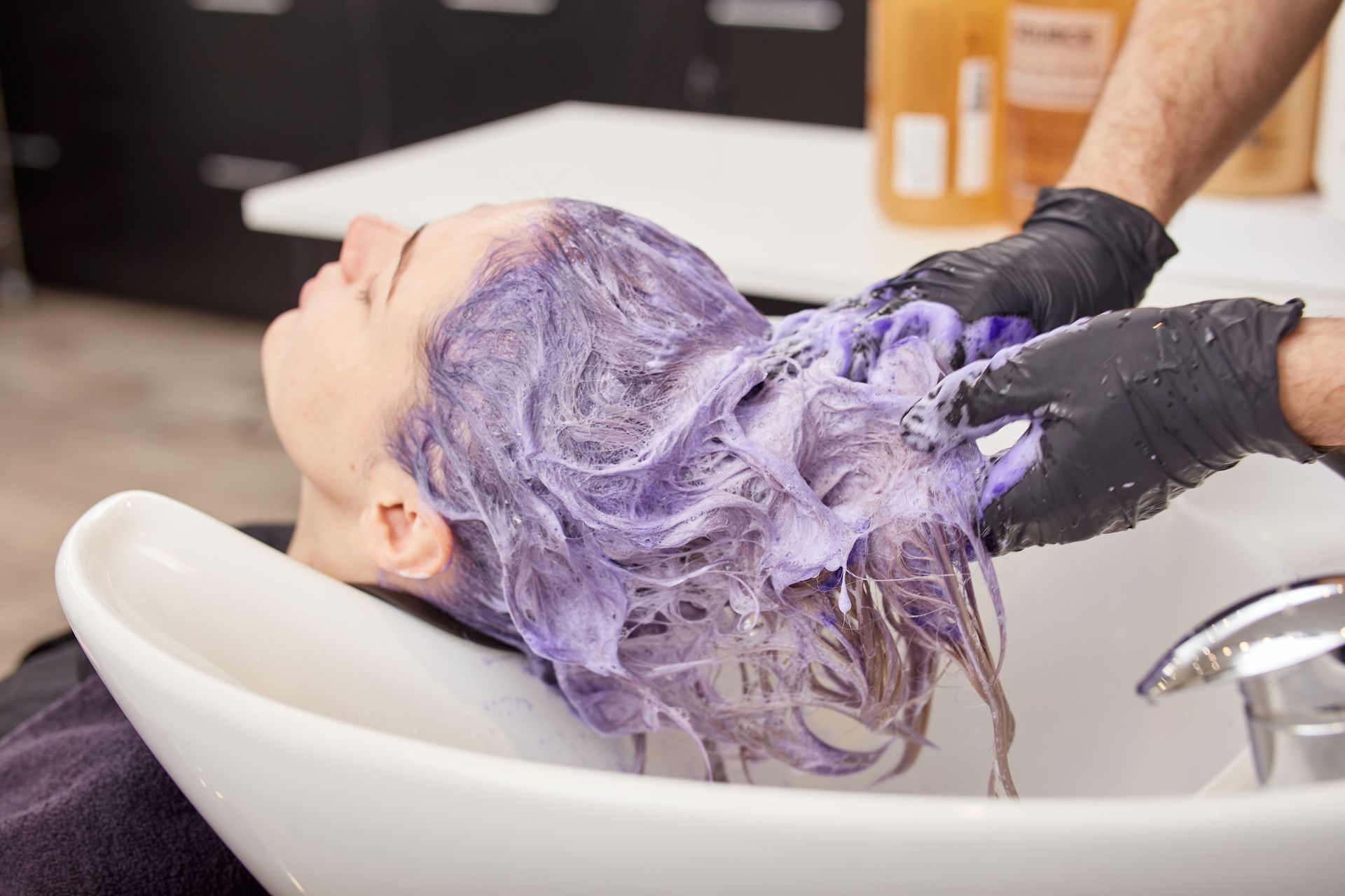 A woman with purple hair is getting her hair washed in a sink.