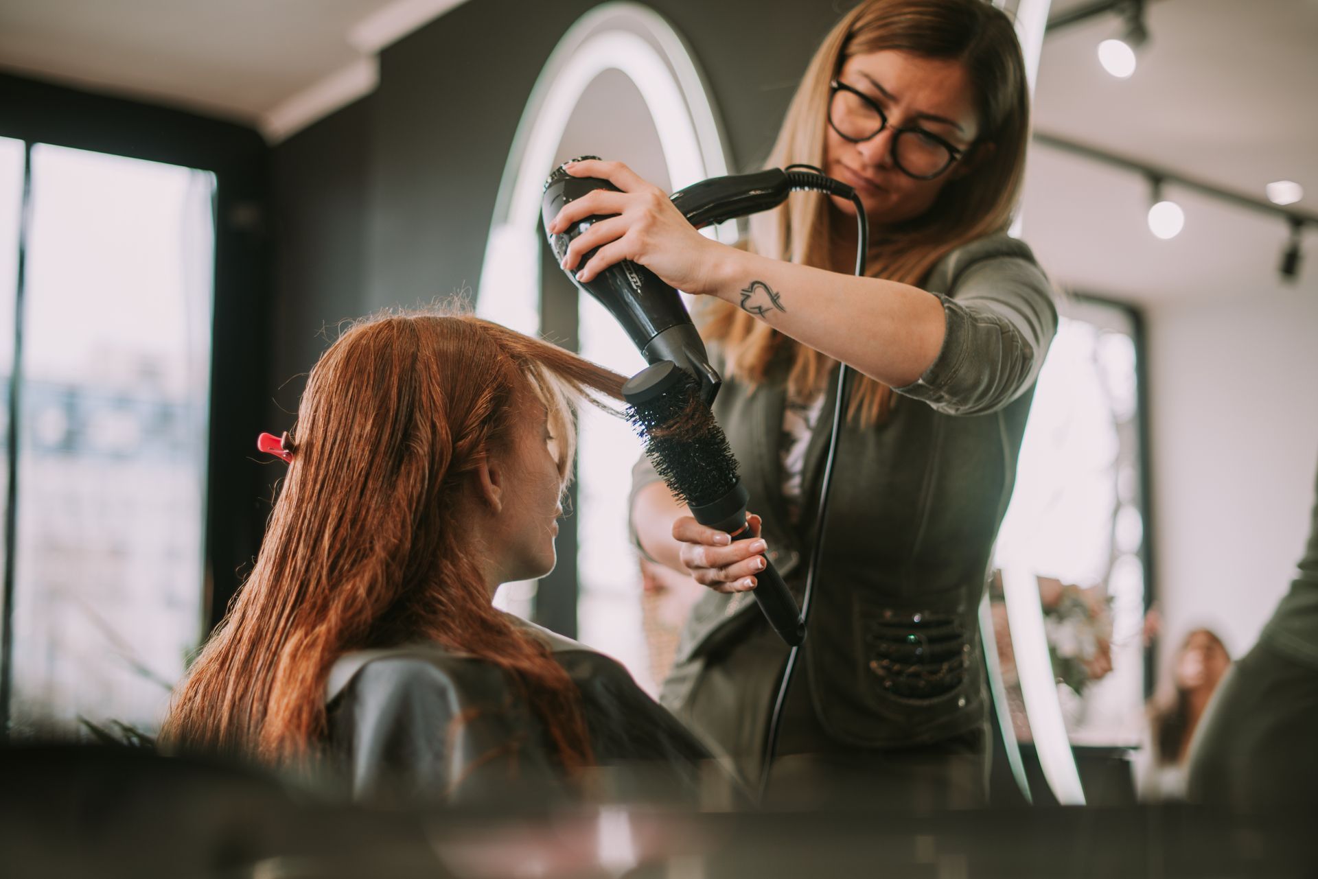 Hairdresser blow-drying a client's red hair in a salon.
