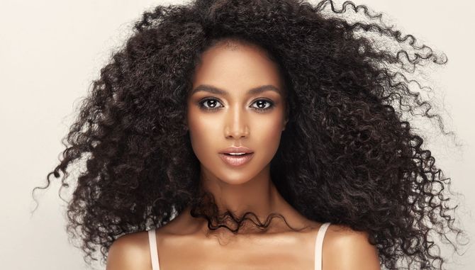 A woman with very long curly hair is wearing a white tank top.