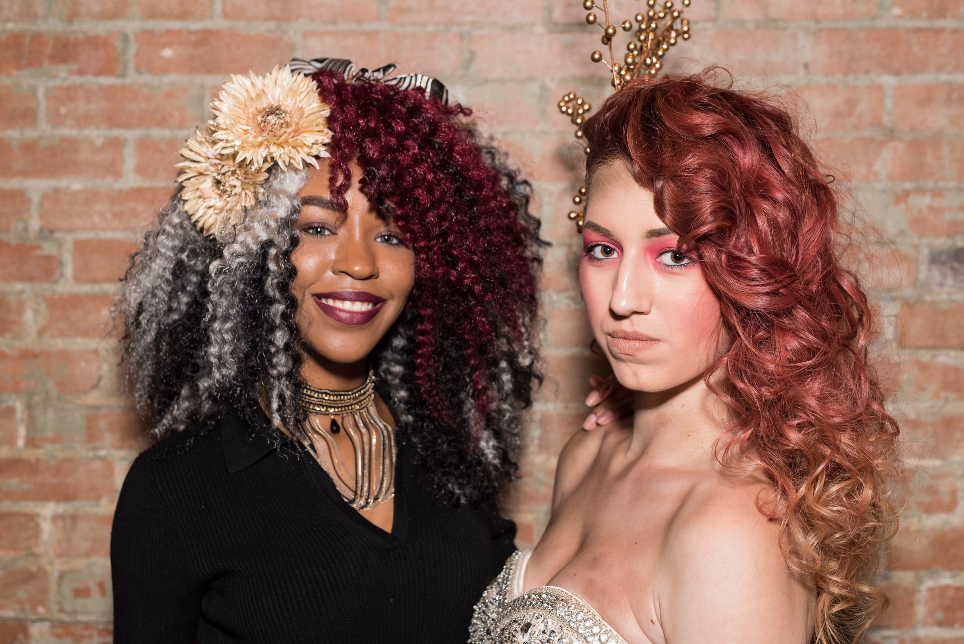 Two people with ornate hairstyles pose in front of a brick wall. One wears black, the other a dress.