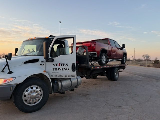 A white States Towing flatbed truck hauling a red pickup truck on a paved road under a clear sky.