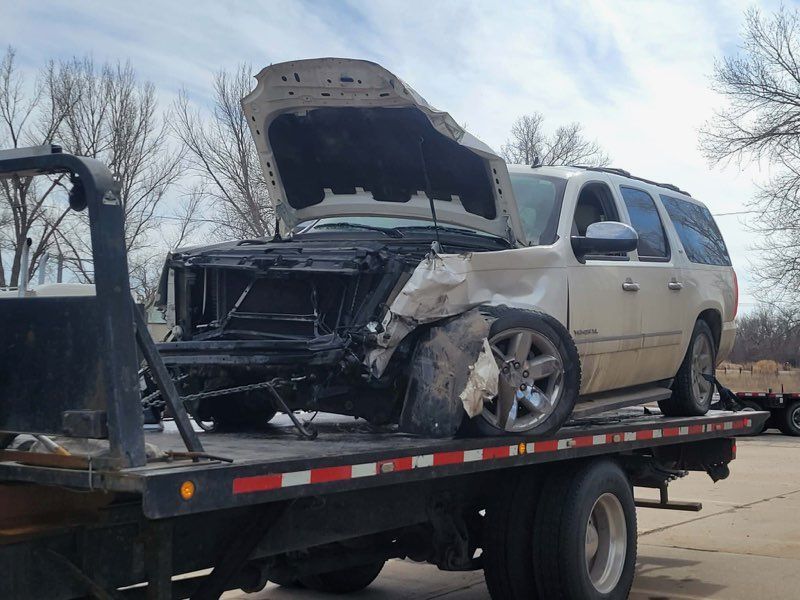 A white SUV with severe front-end damage and an open hood secured on the bed of a flatbed tow truck.