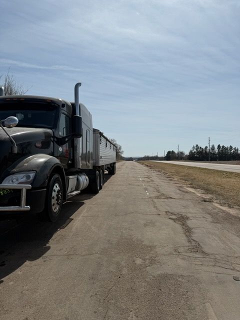A gray semi-truck parked on the cracked pavement of a roadside shoulder next to an open highway under a sunny sky.