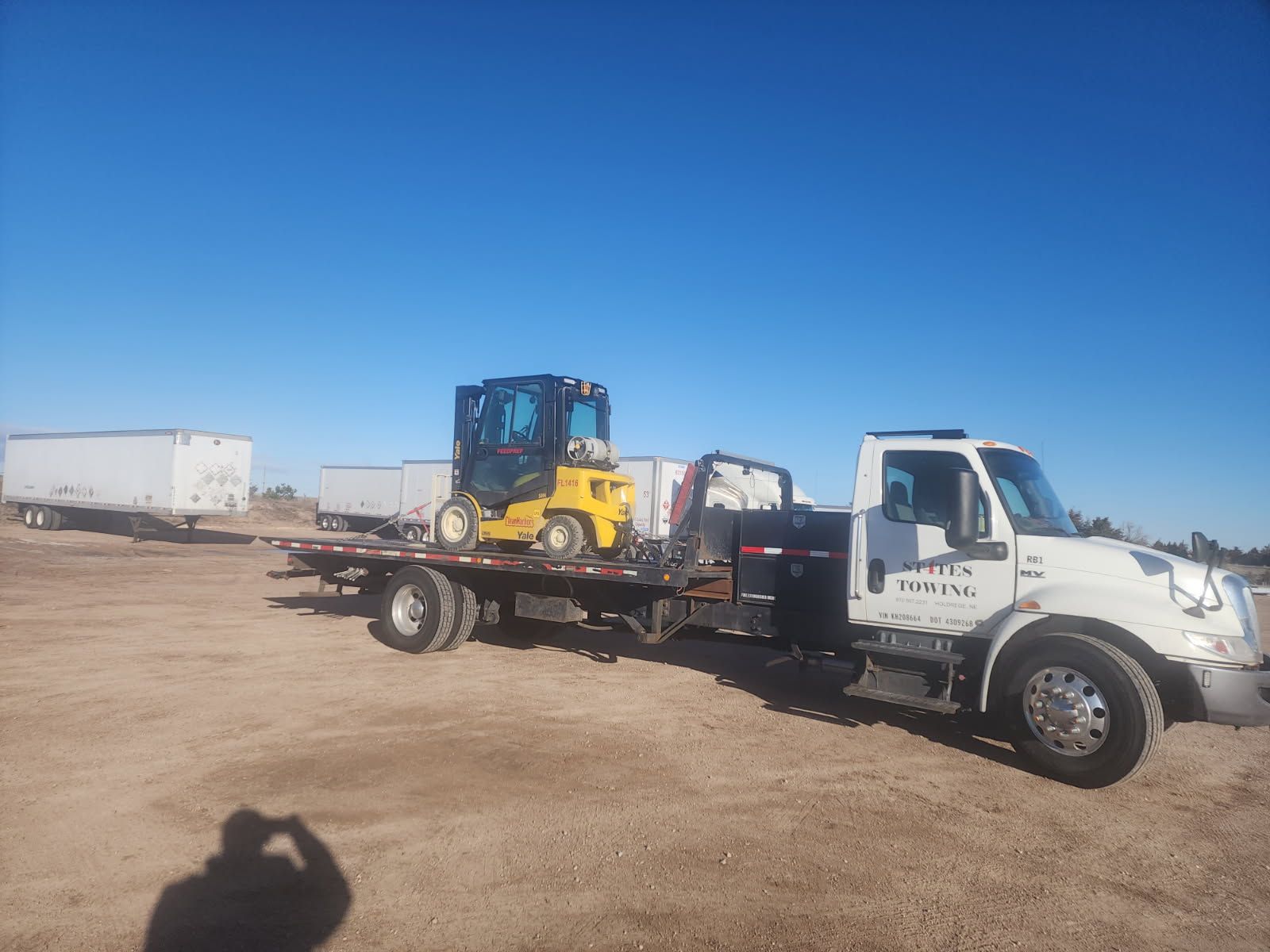 A white flatbed truck carrying a yellow compact excavator parked on a dirt lot under a clear blue sky.