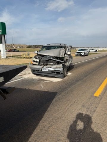 A heavily damaged silver SUV sits on the shoulder of a highway, with a tow truck ramp visible in the foreground.