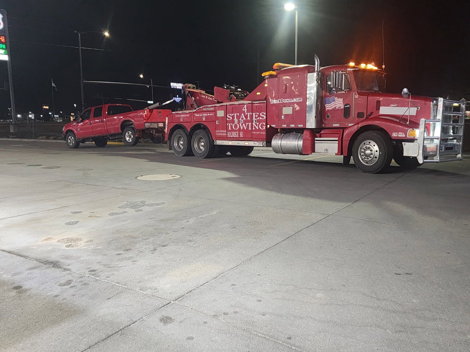 A red tow truck pulling a red pickup truck at a gas station parking lot at night.