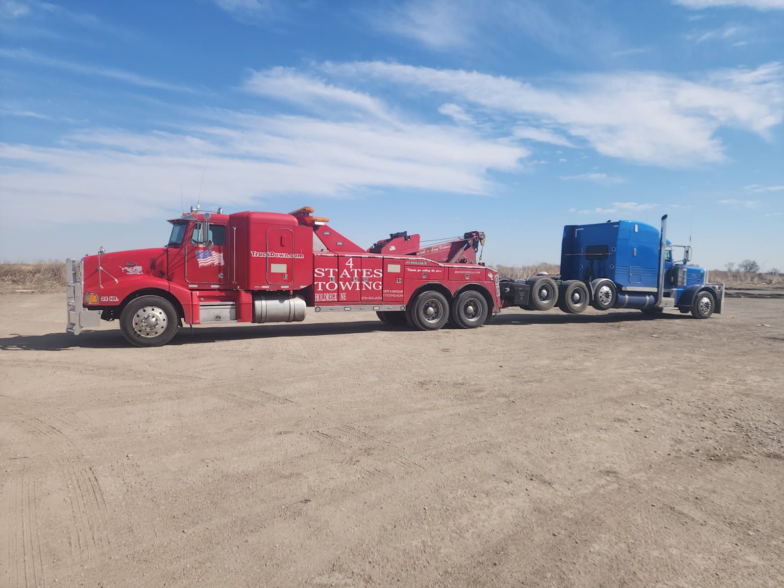 A red tow truck is towing a blue semi-truck on a gravel lot under a clear blue sky.