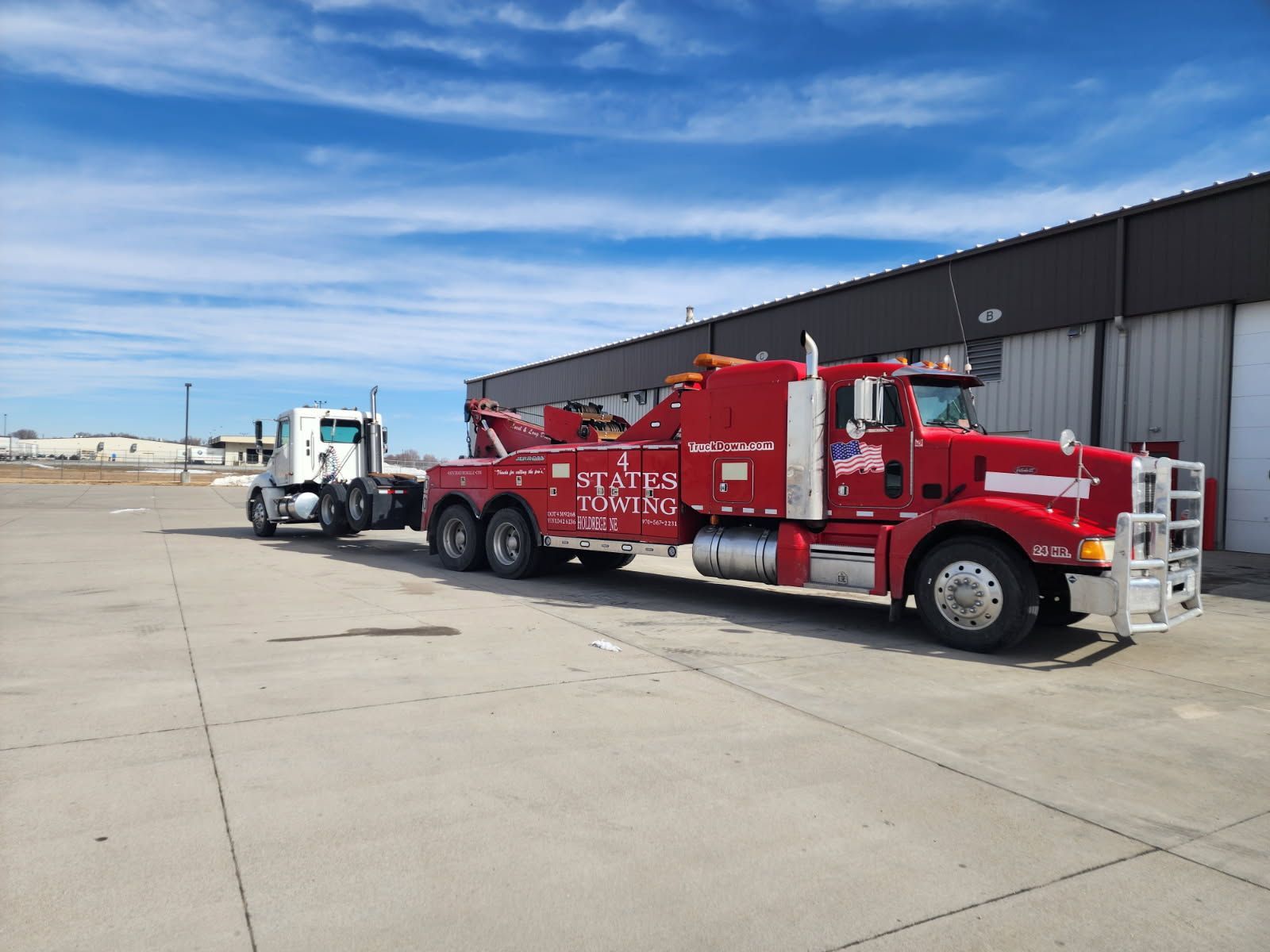 A red tow truck pulling a white semi-truck tractor on an asphalt lot in front of a long building under a blue sky.