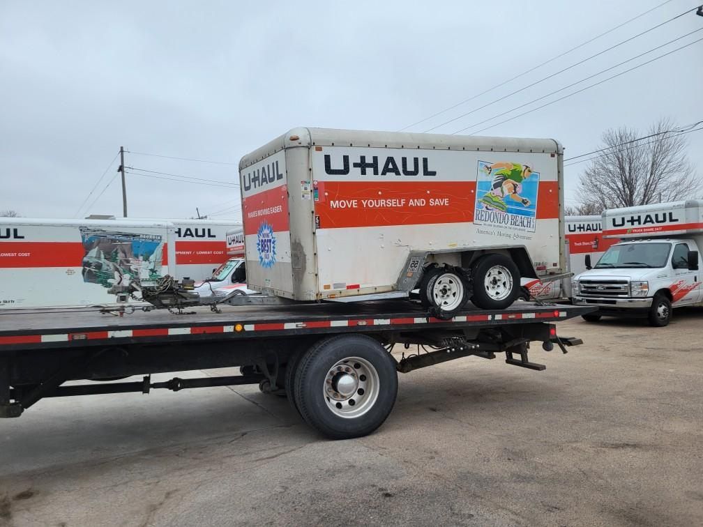 A U-Haul cargo trailer is secured on a flatbed trailer in a parking lot, with other U-Haul vehicles visible nearby.
