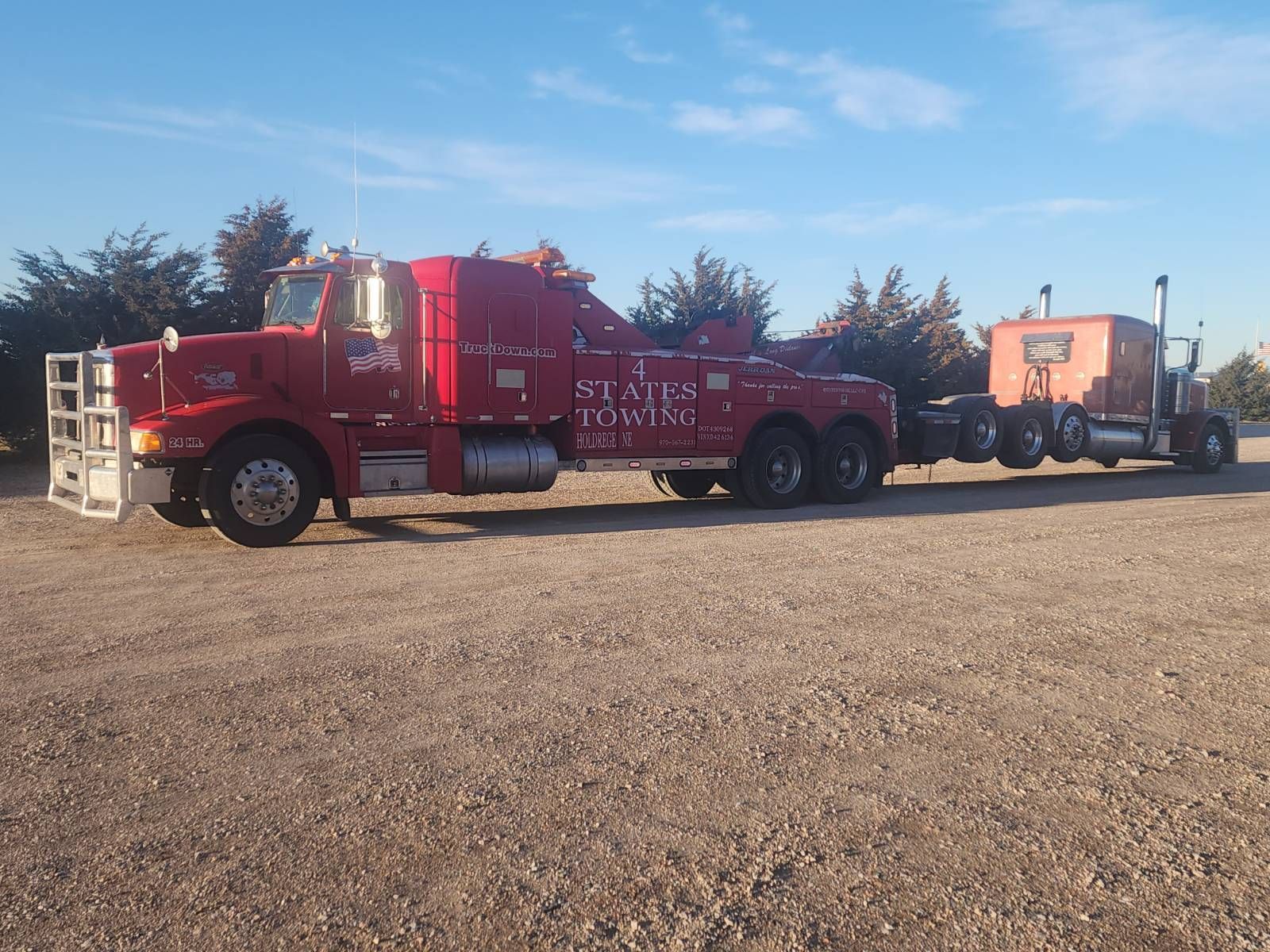 A red heavy-duty tow truck is parked on a gravel lot, towing another semi-truck cab behind it under a blue sky.