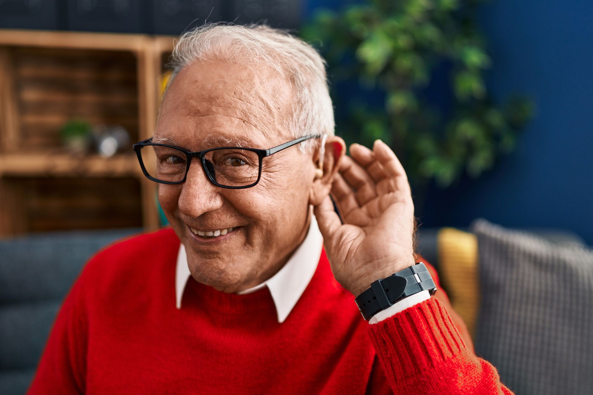 An elderly man wearing glasses and a red sweater is listening to something.