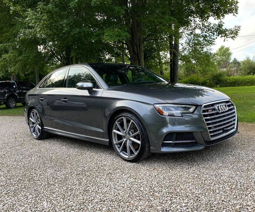 Gray Audi sedan parked on gravel, with trees in the background.