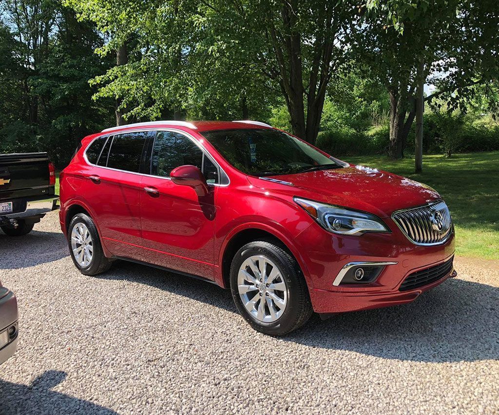 Red Buick Envision SUV parked on gravel driveway. Trees and grassy area in background. Sunny day.