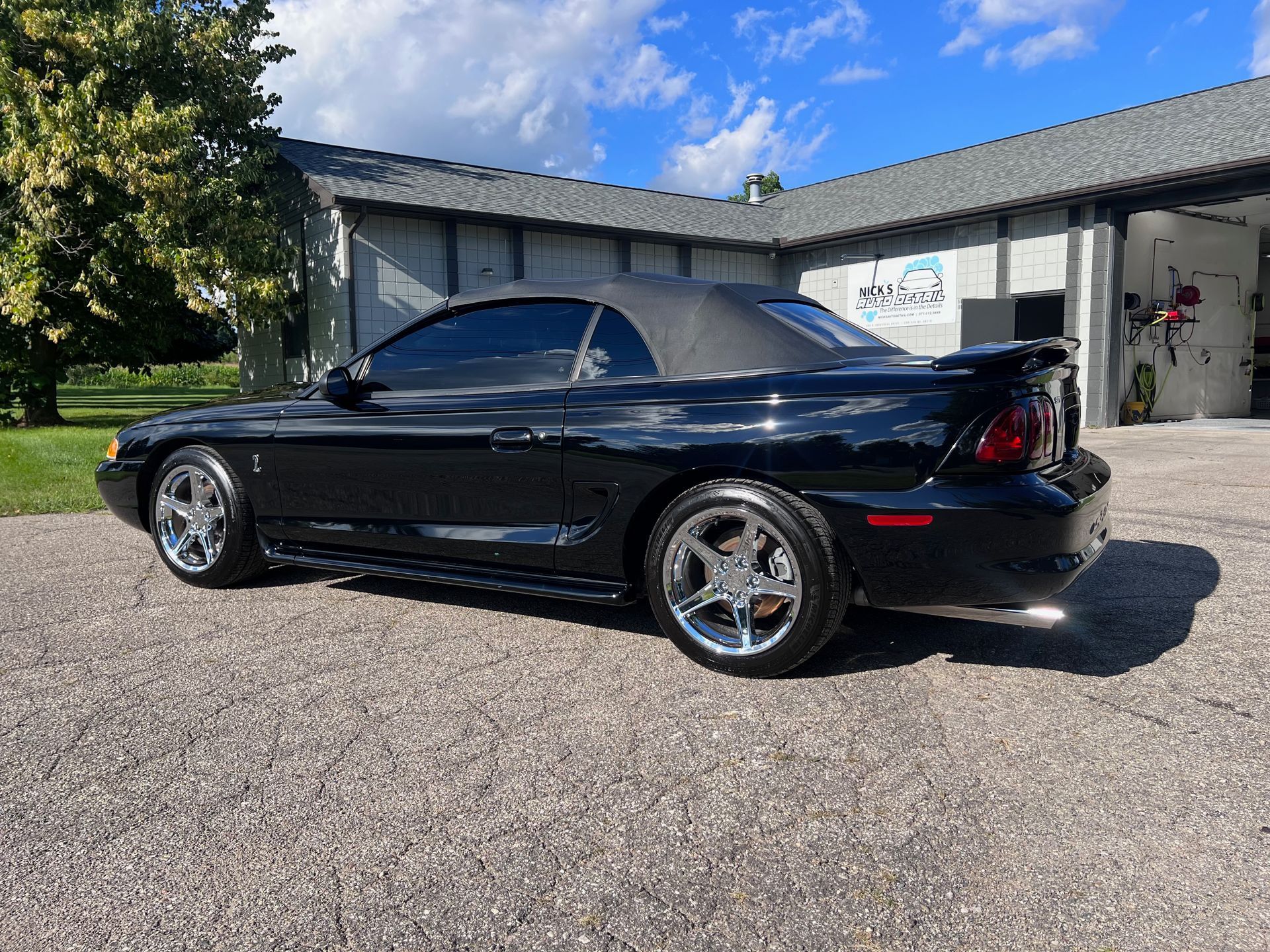 Black Ford Mustang convertible, chrome wheels, parked in front of a building with a blue sky.