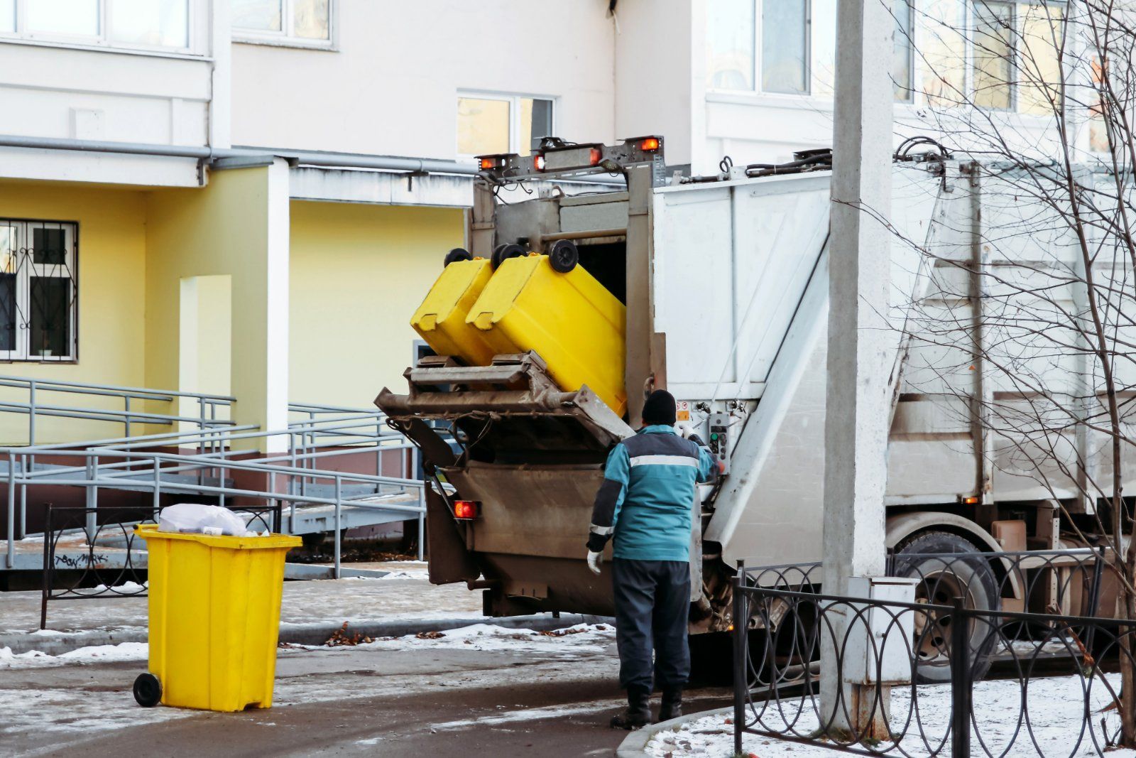 A worker loads yellow dumpsters into the back of a garbage truck parked in front of a building.