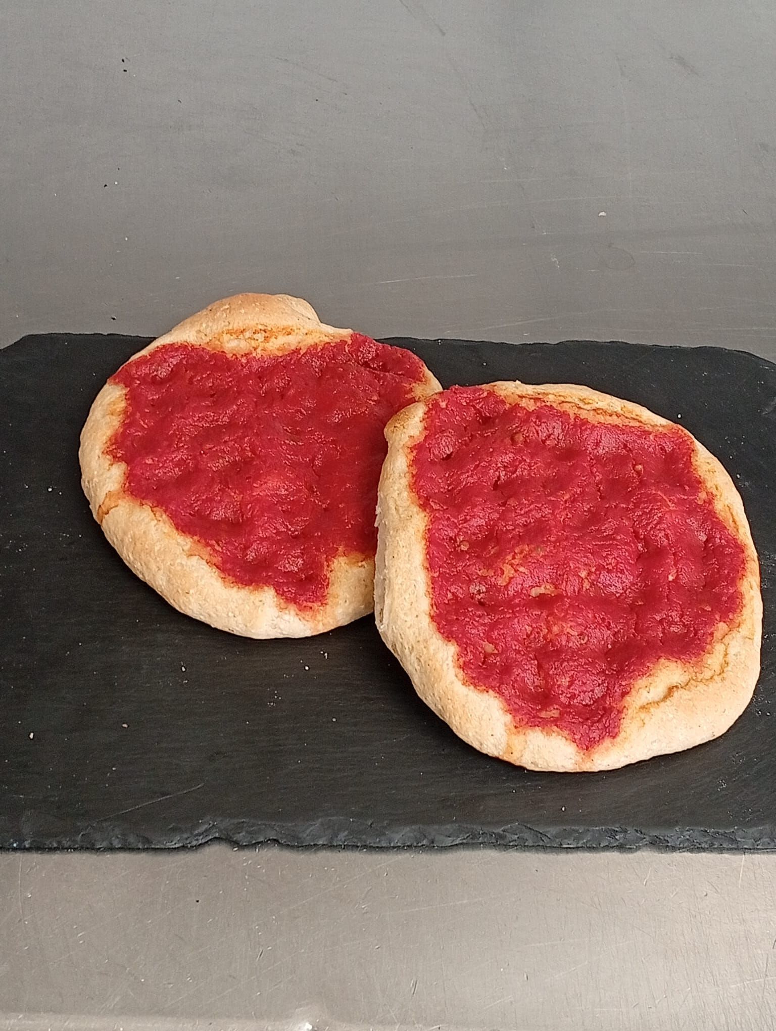 Due focaccine rotonde, simili a focacce, farcite con marmellata rossa, disposte su un tagliere rettangolare di pietra nera.