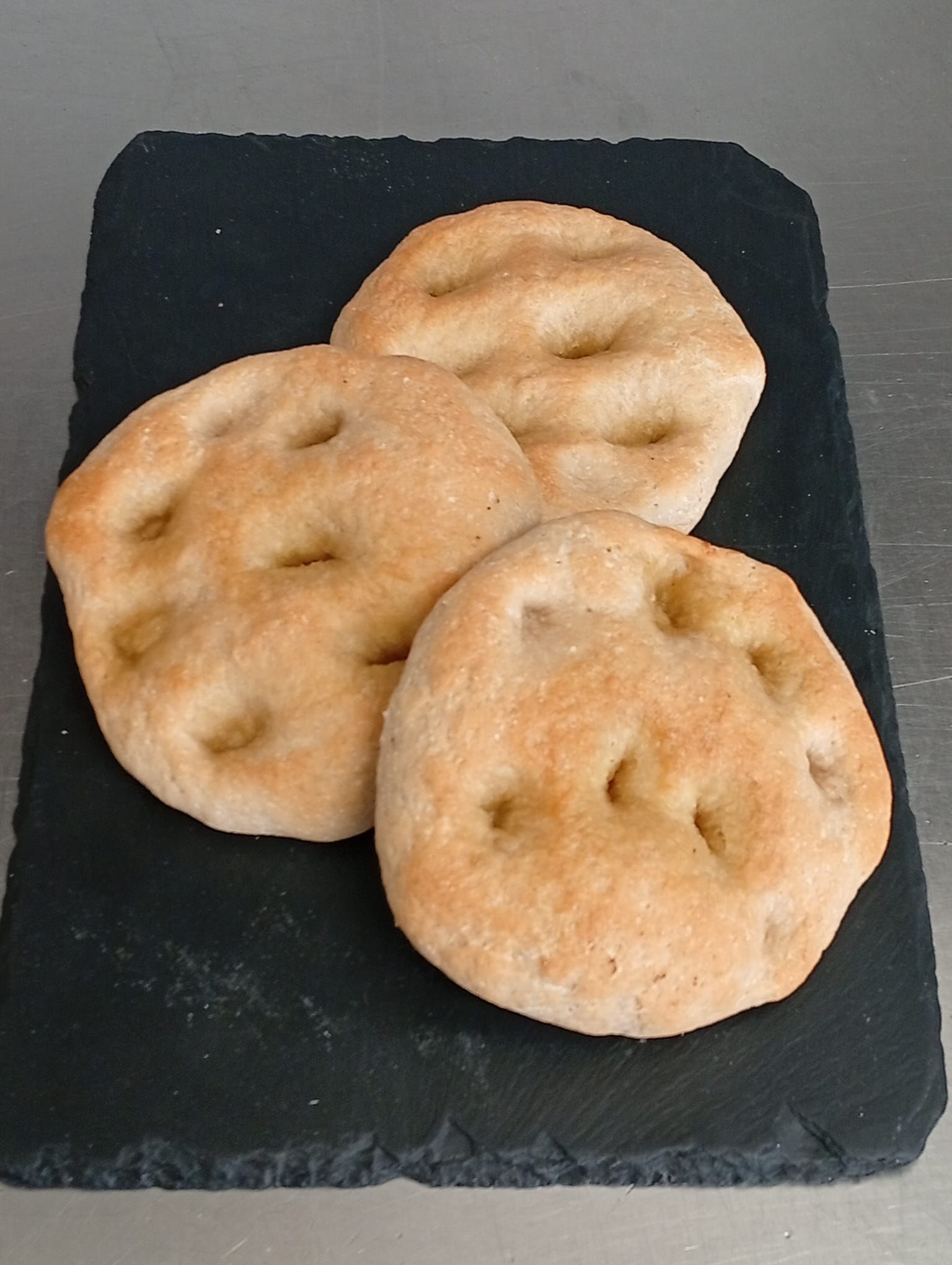 Tre piccole focacce rotonde con le caratteristiche fossette, disposte su un tagliere rettangolare di ardesia nera.