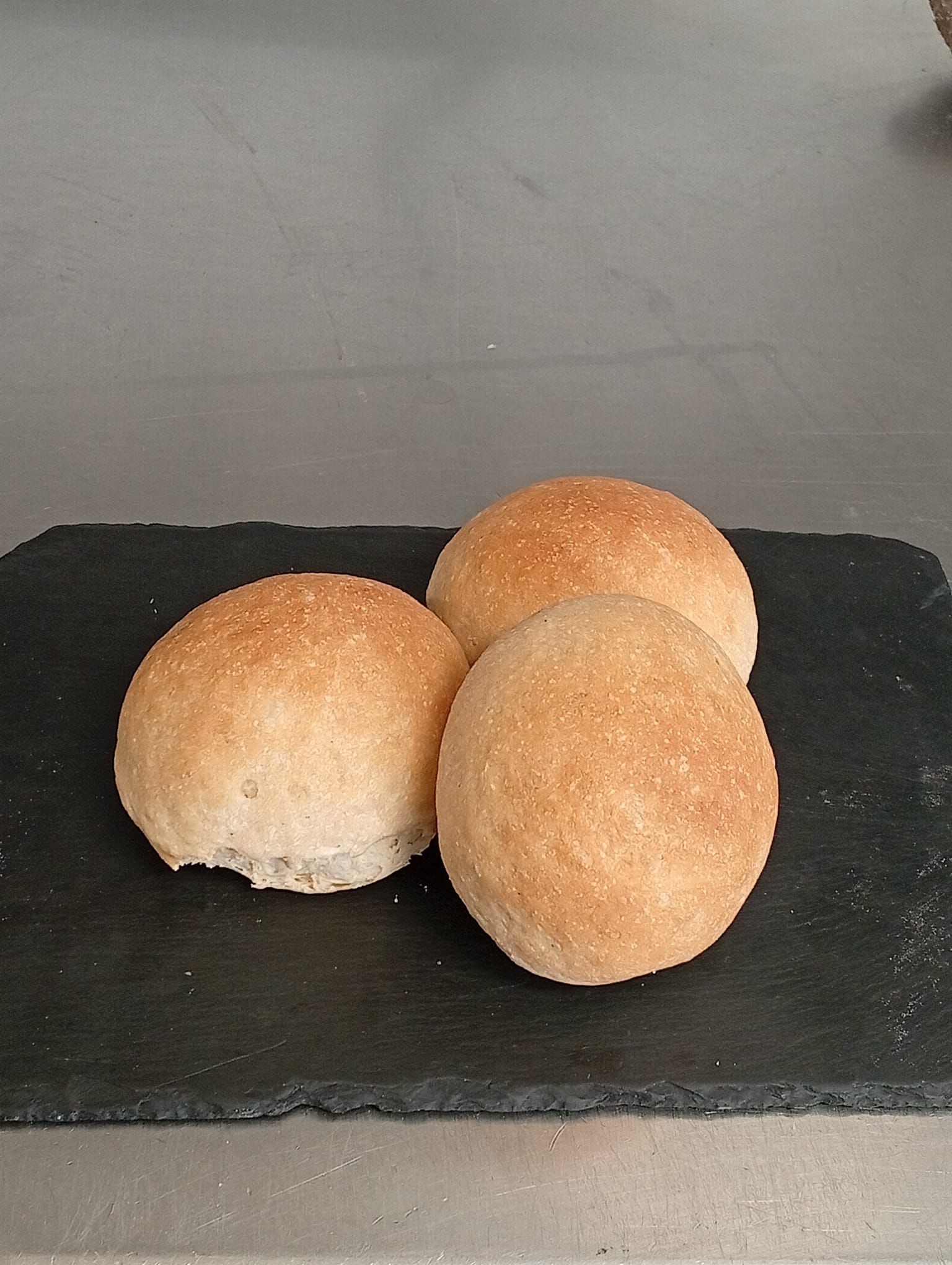 Tre panini rotondi di colore marrone dorato disposti su un tagliere rettangolare di ardesia nera.