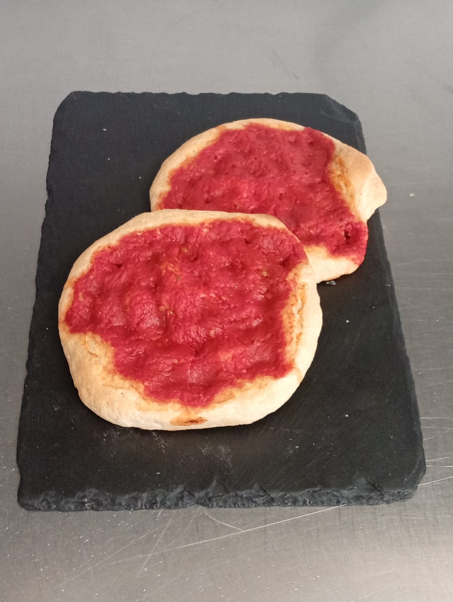 Due piccole focacce rotonde condite con salsa di pomodoro rosso, adagiate su un tagliere rettangolare di ardesia nera.