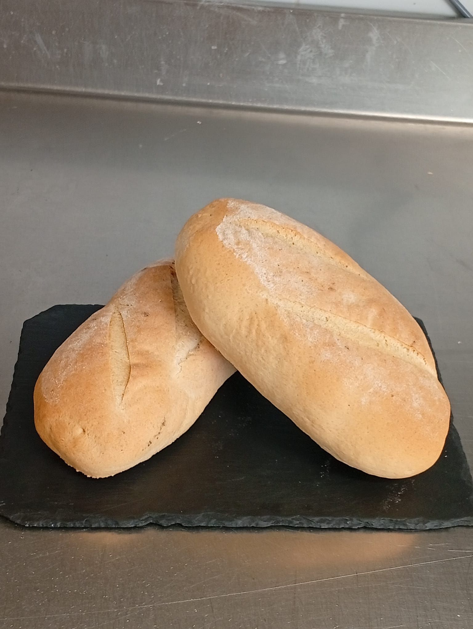 Due panini croccanti e dorati, adagiati su un tagliere rettangolare di ardesia nera, su uno sfondo di acciaio inossidabile.