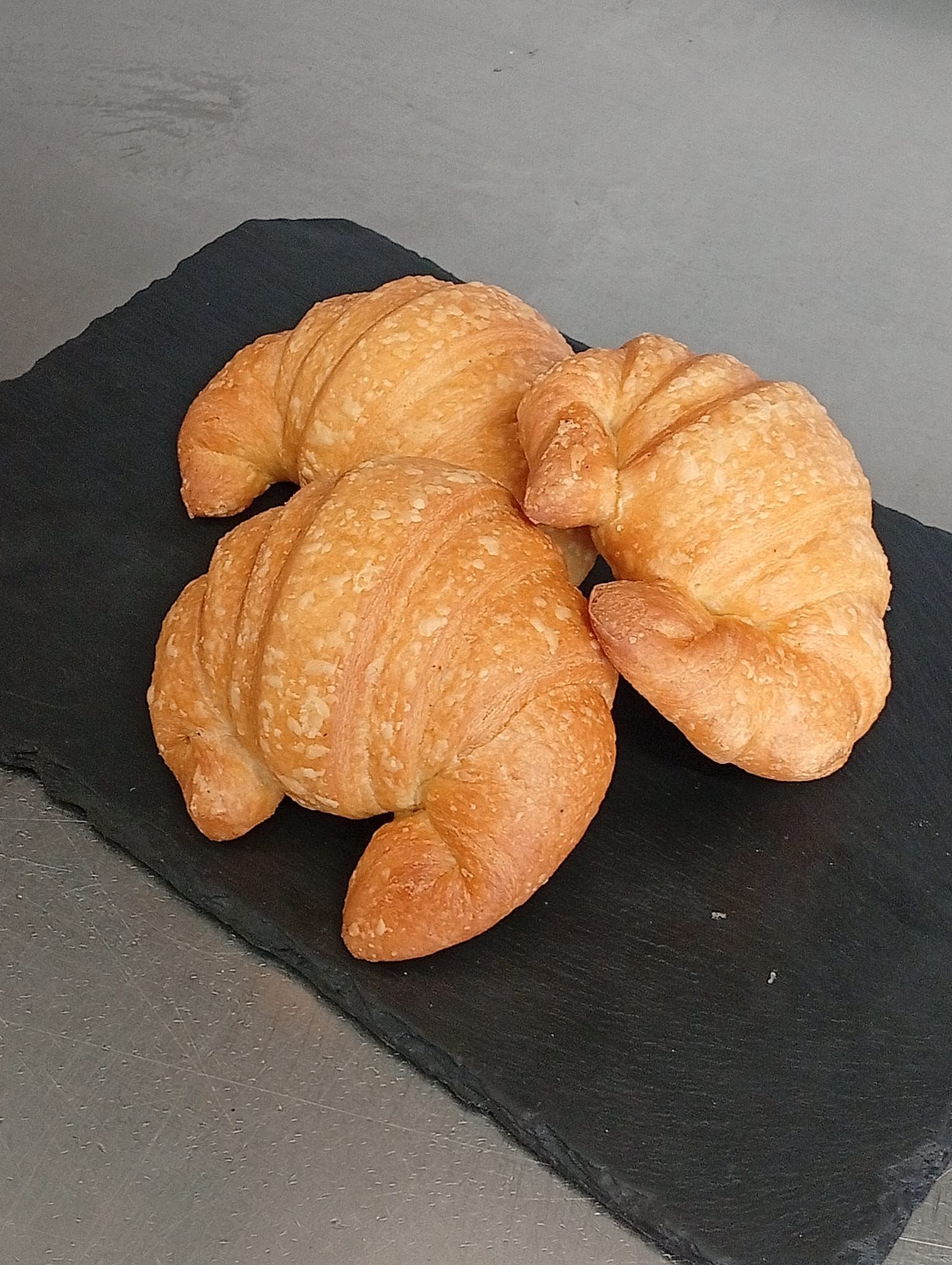 Tre croissant dorati disposti su un piatto da portata rettangolare in pietra scura, su uno sfondo neutro.