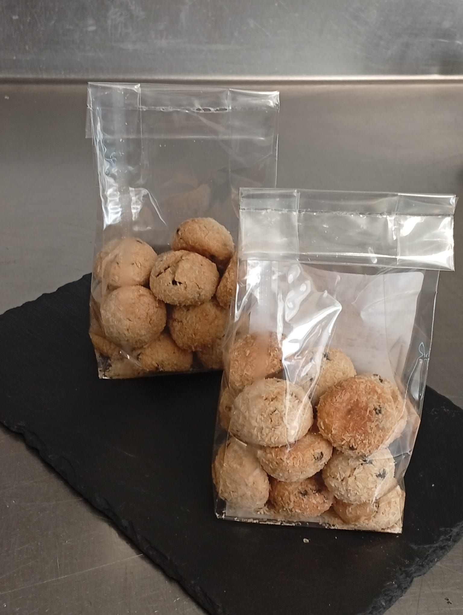 Due sacchetti di plastica trasparente pieni di biscotti rotondi color marrone dorato, appoggiati su una lastra di ardesia